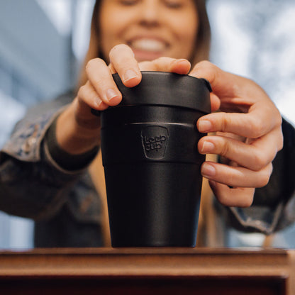 Woman holding black reusable coffee cup with lid on wooden table, smiling in blurred background