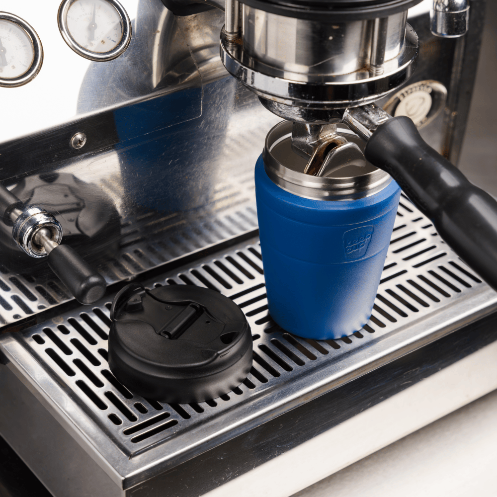 KeepCup Commuter M 12oz in Cobalt Black positioned under an espresso machine group head, with the black flip lid removed and placed on the stainless steel drip tray beside it.