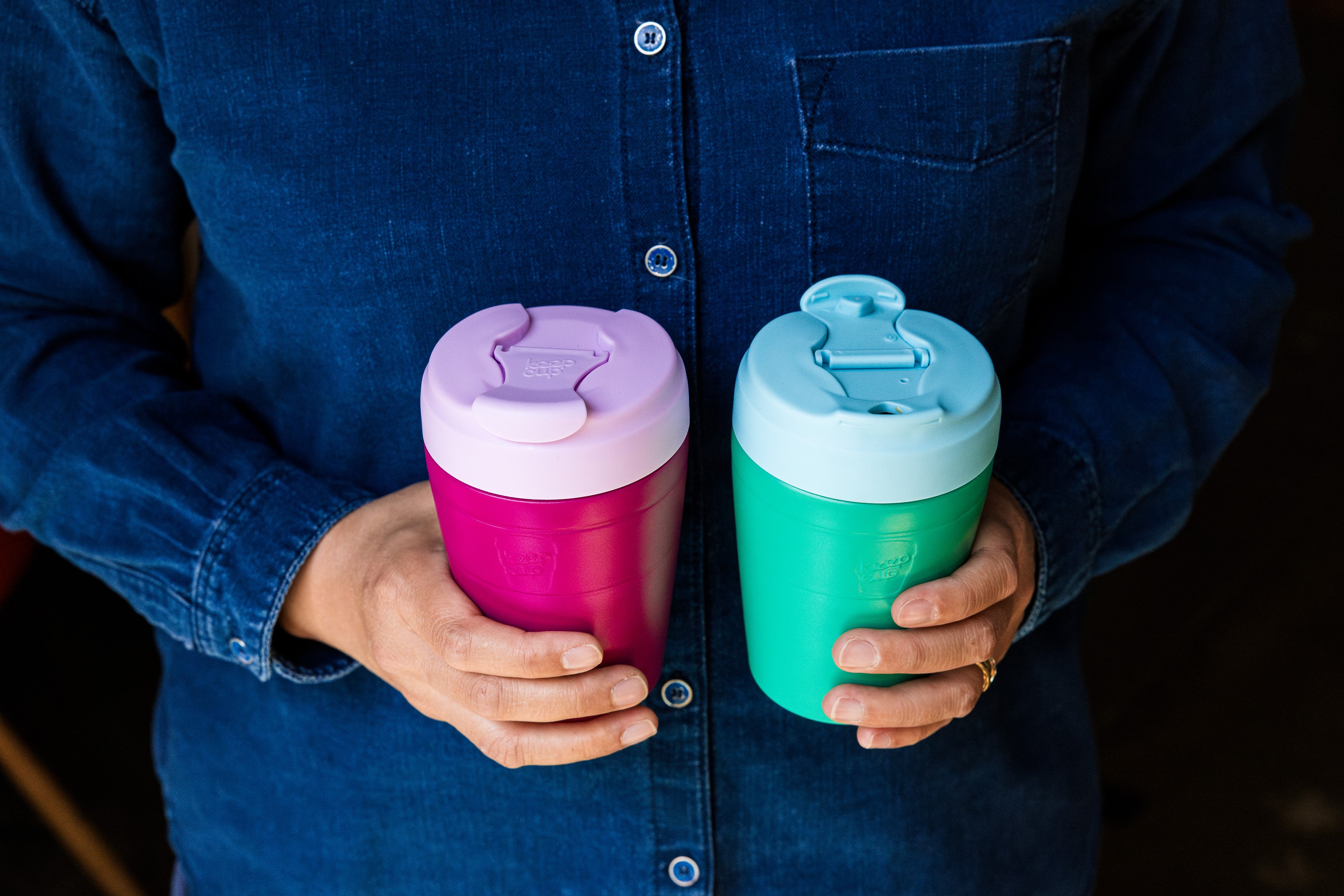 Person holding two colorful tumblers with lids against a dark background