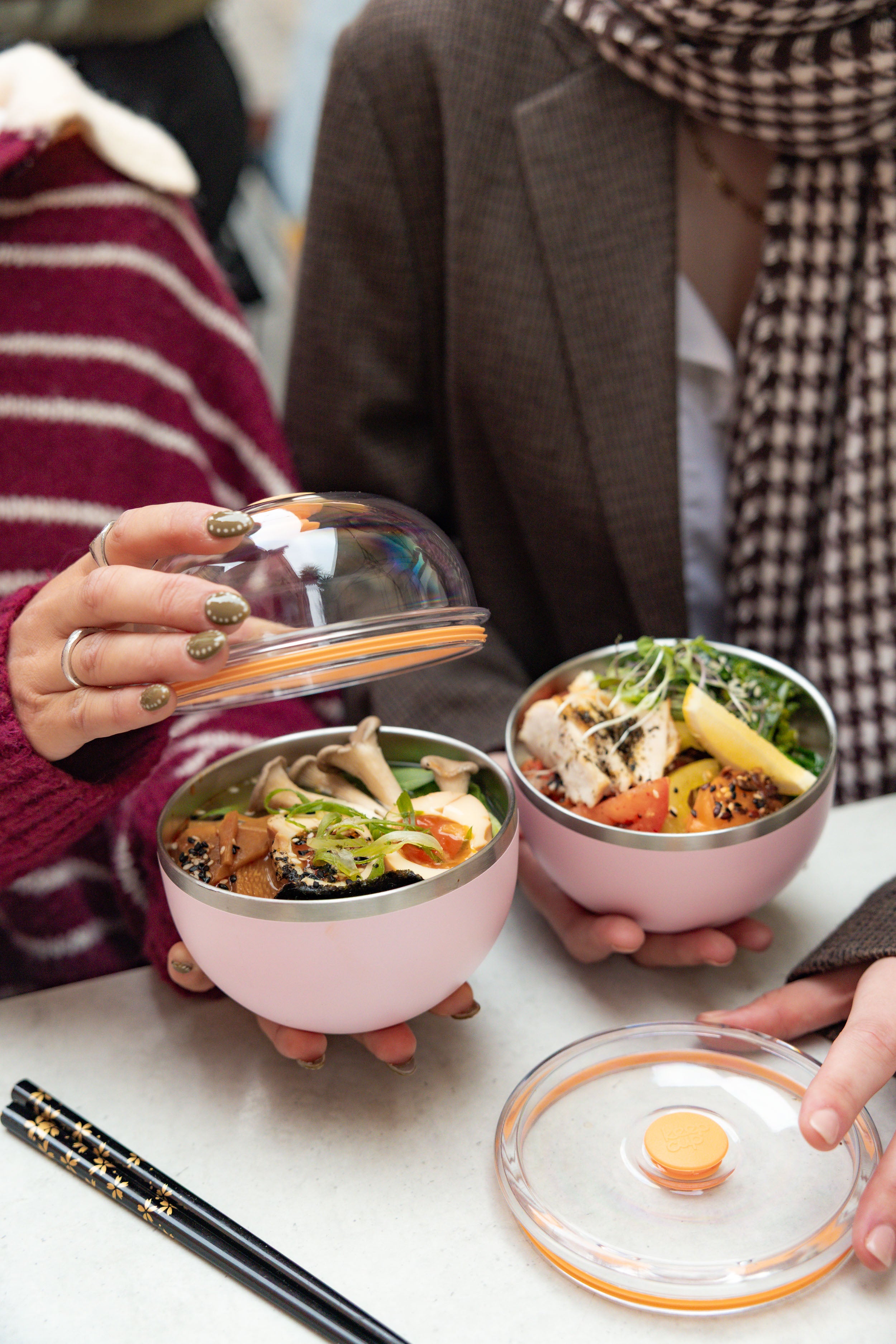 Two people opening Go Bowl Luxe containers in Pashmak Pink, revealing Asian-inspired meals with tofu, mushrooms, and vegetables inside the stainless steel bowls, with clear domed lids with orange seals and black chopsticks on a white table.