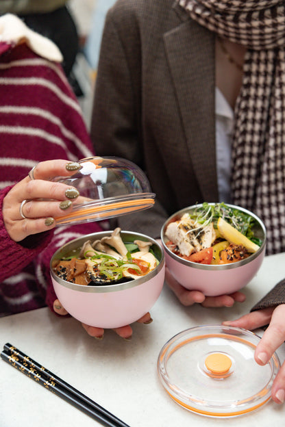 Two people opening Go Bowl Luxe containers in Pashmak Pink, revealing Asian-inspired meals with tofu, mushrooms, and vegetables inside the stainless steel bowls, with clear domed lids with orange seals and black chopsticks on a white table.