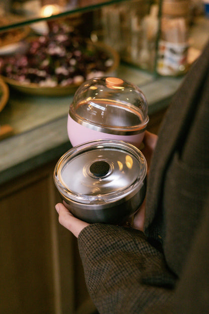 A hand holds two KeepCup Go Bowl products in a café setting - the Go Bowl Luxe in Pashmak Pink with a domed lid and peach button on top, alongside a Nitro bowl with a flat lid featuring a black center button.