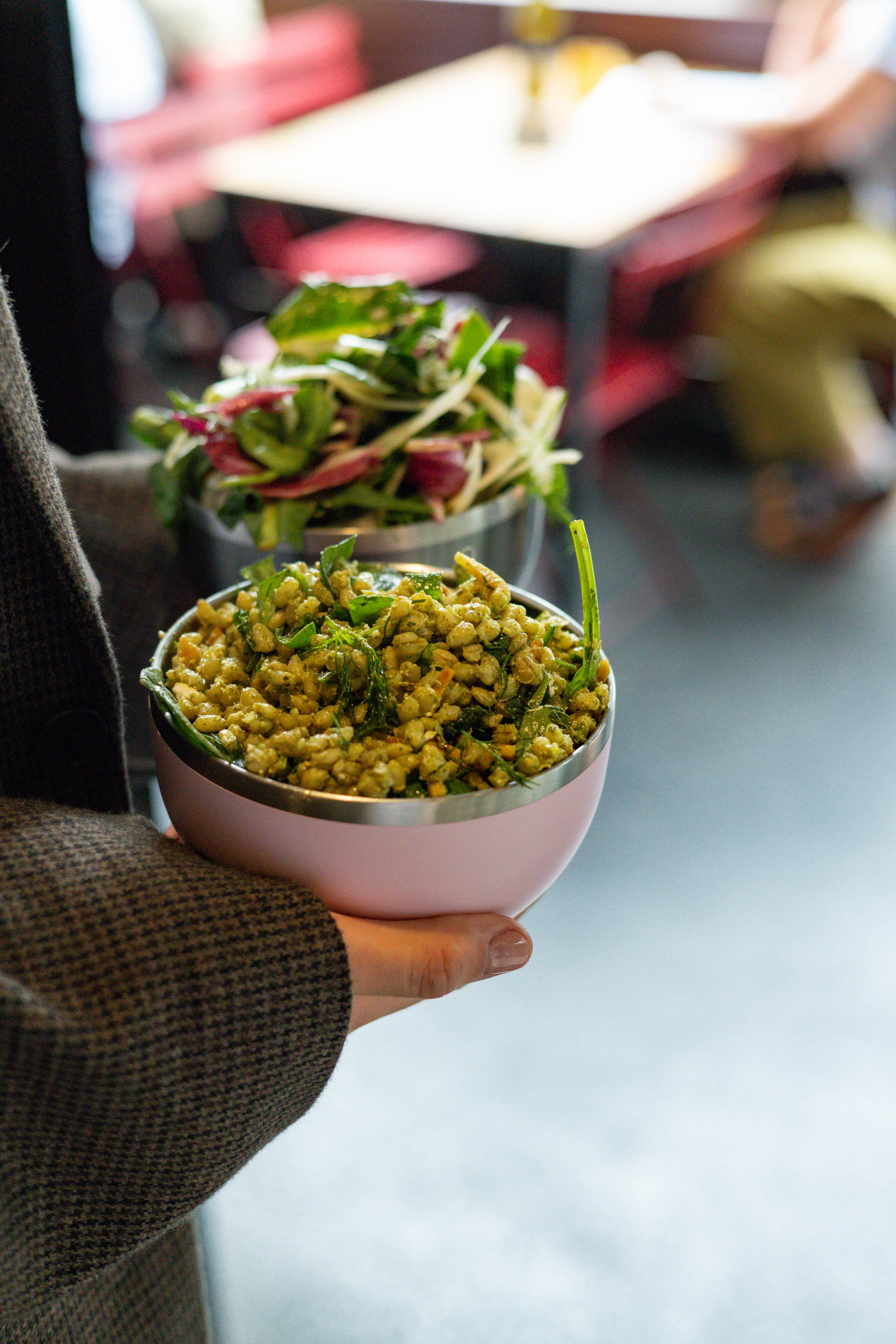 Hand holding a Go Bowl Luxe in Pashmak Pink filled with a grain salad topped with fresh herbs, with a second bowl containing mixed greens visible in the background at a casual dining setting.