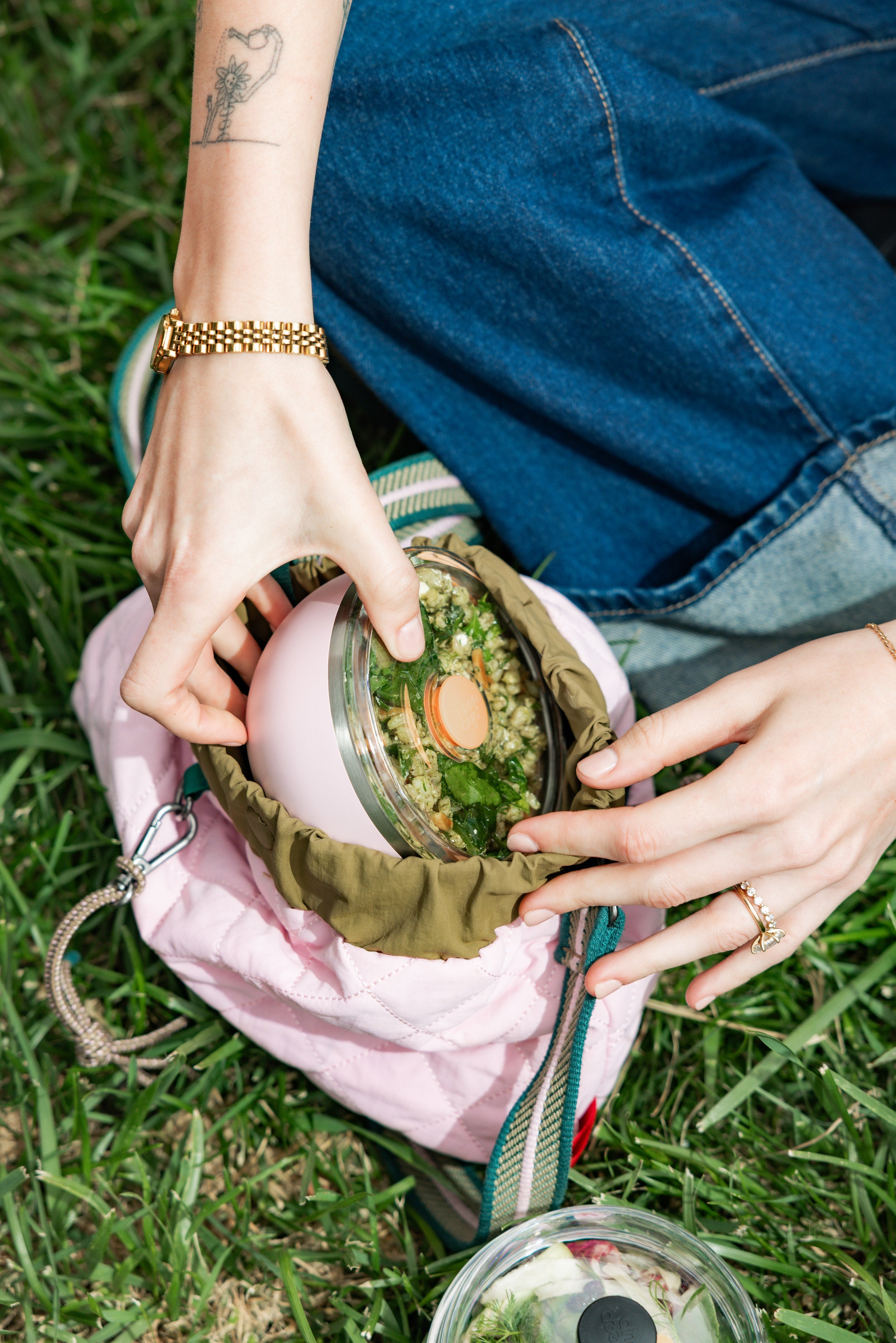 Hands reaching into a pink quilted lunch bag to retrieve a KeepCup Go Bowl Luxe in Pashmak Pink with flat lid, containing a green grain salad, set on grass during an outdoor picnic.