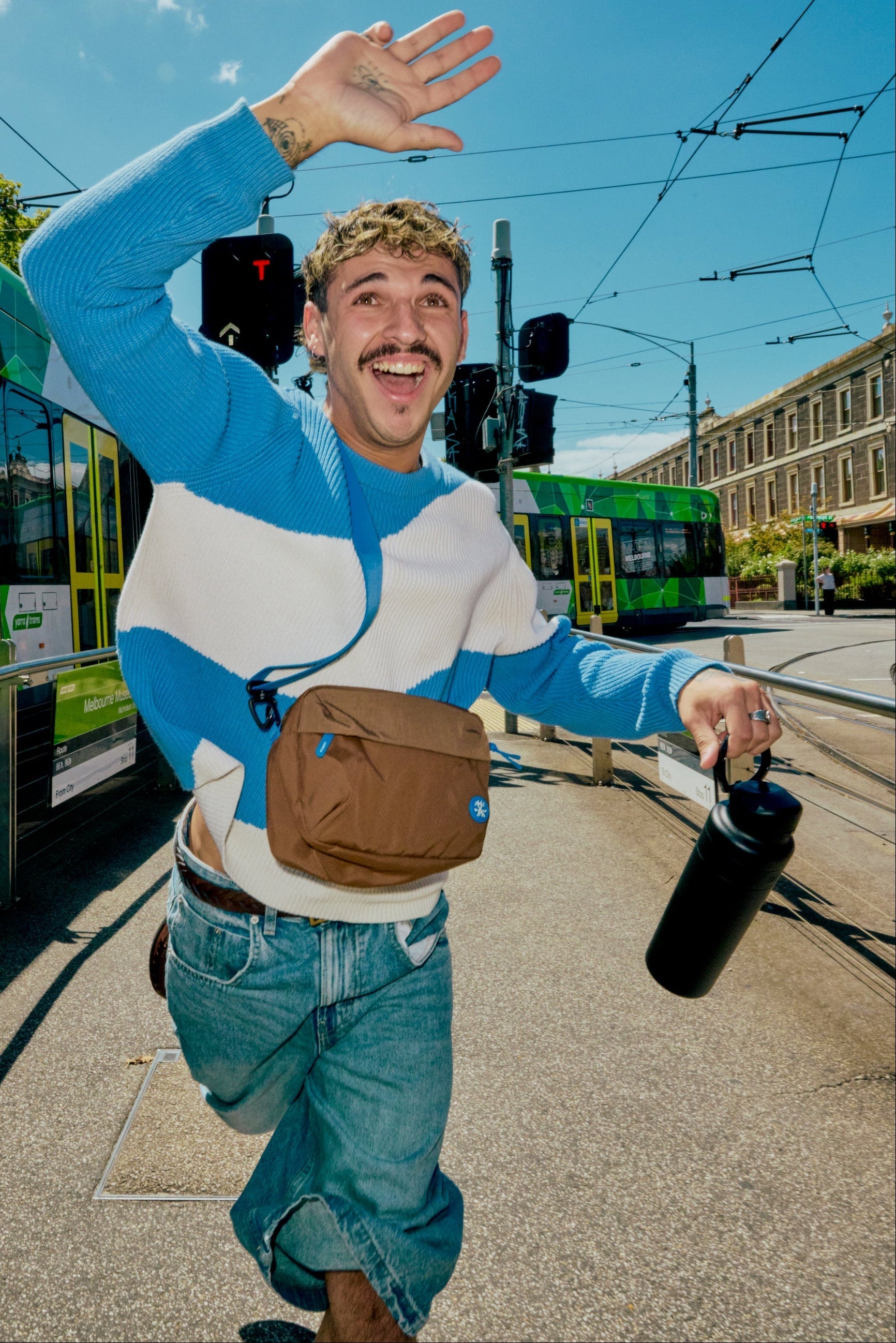 Man in a blue and white sweater with a brown bag and black bottle on a street with trams in the background.
