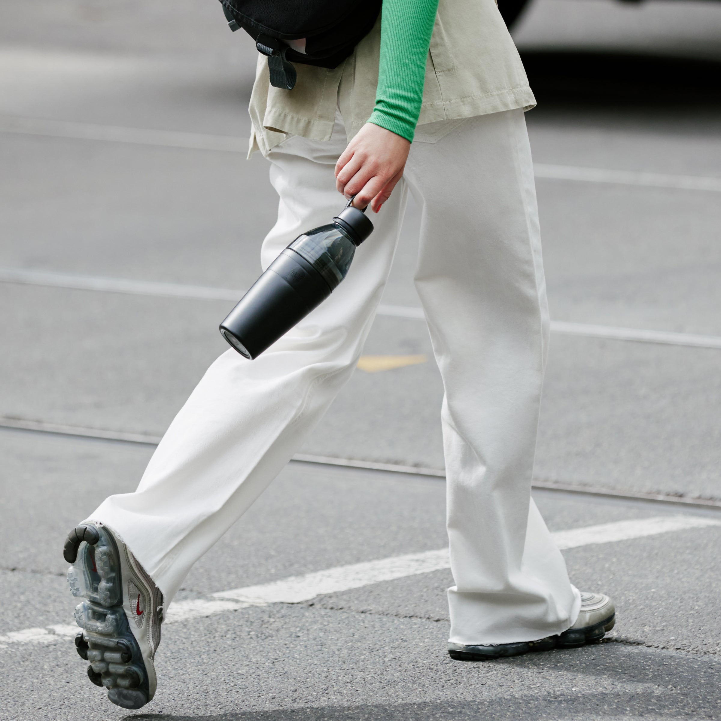 Person walking on a street holding a black KeepCup water bottle.