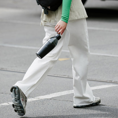 Person walking on a street holding a black KeepCup water bottle.