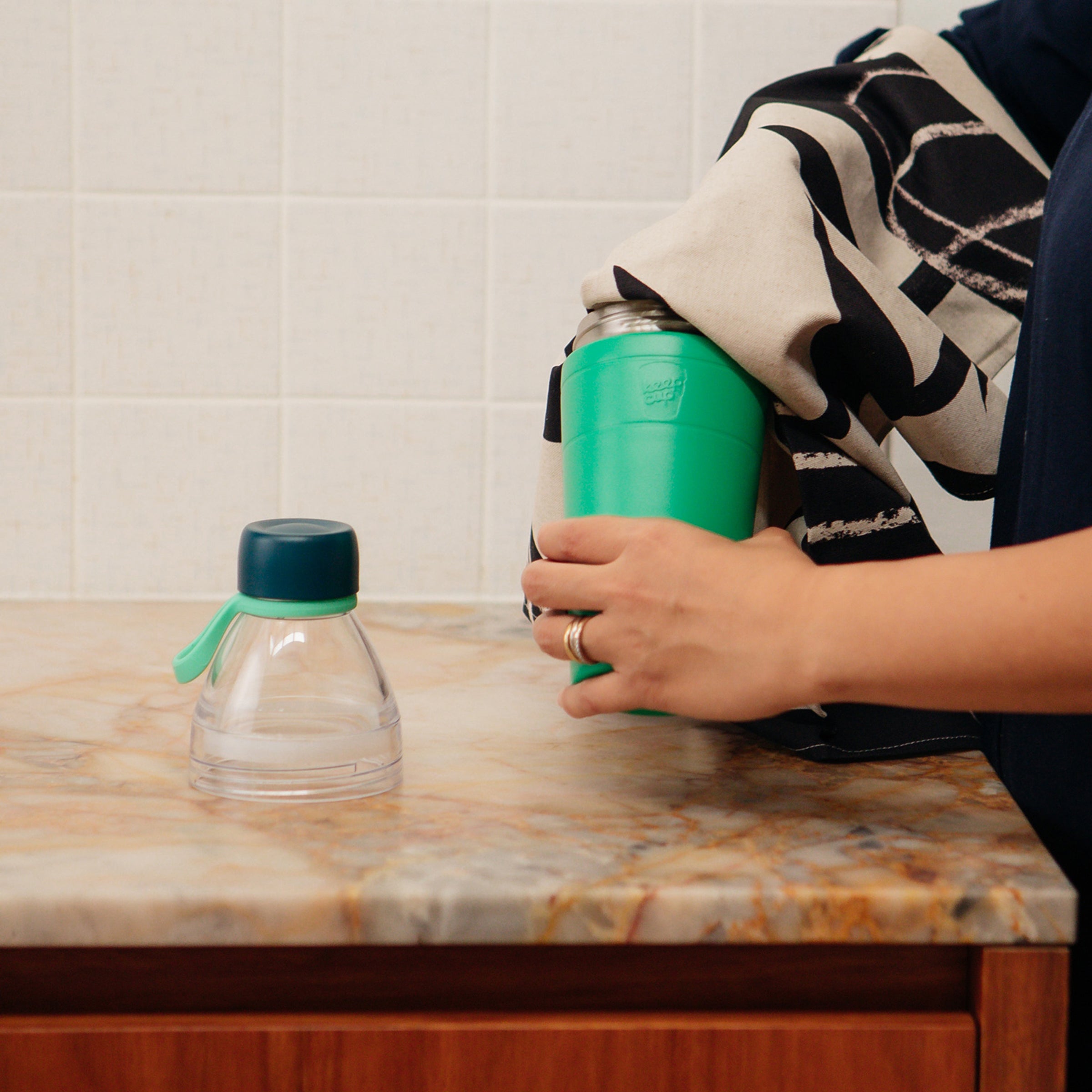 Person holding and cleaning a KeepCup water bottle on a marble countertop with a tiled wall background
