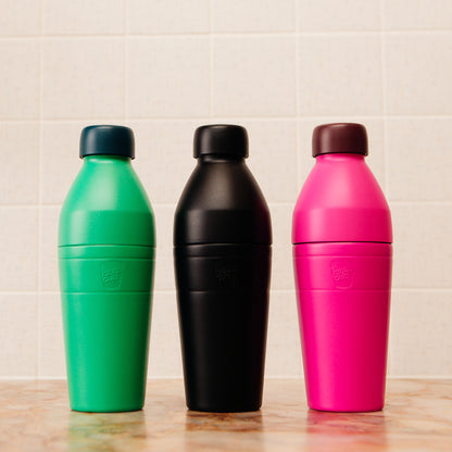 Three colorful KeepCup insulated water bottles (green, black, pink) on a wooden surface with a tiled wall background.