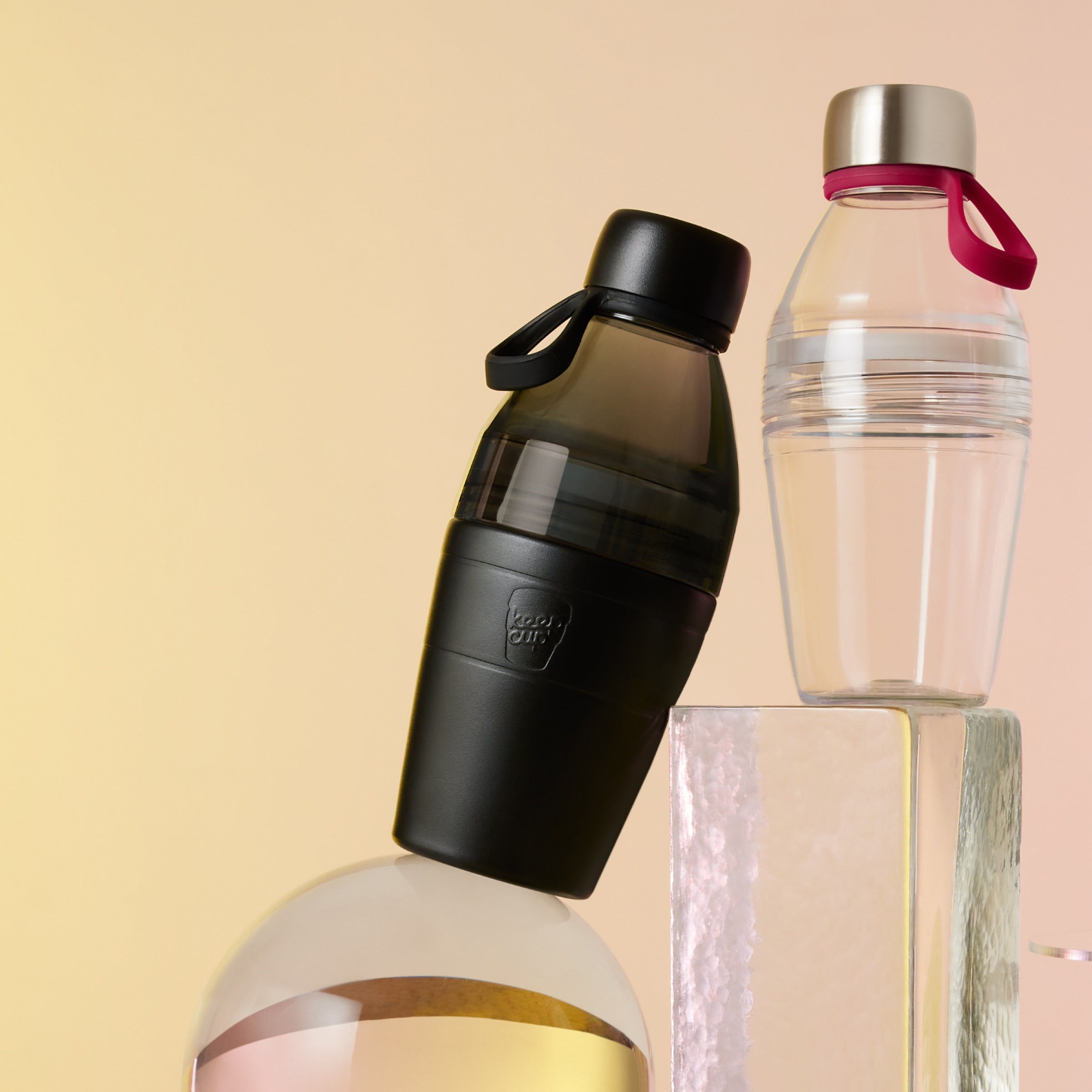 Two KeepCup water bottles, one black and one clear with a pink carry handle, on a beige studio shot background.