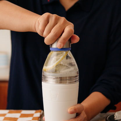 Person opening a white KeepCup water bottle with a lemon slice inside, kitchen setting with blurred background