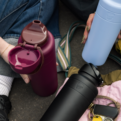 A hand holds the open KeepCup Ora Bottle 24oz in Mulberry, showing its flip-top lid and drinking spout, alongside a light blue and black KeepCup bottle, all resting on colorful bags and accessories in a casual outdoor setting.