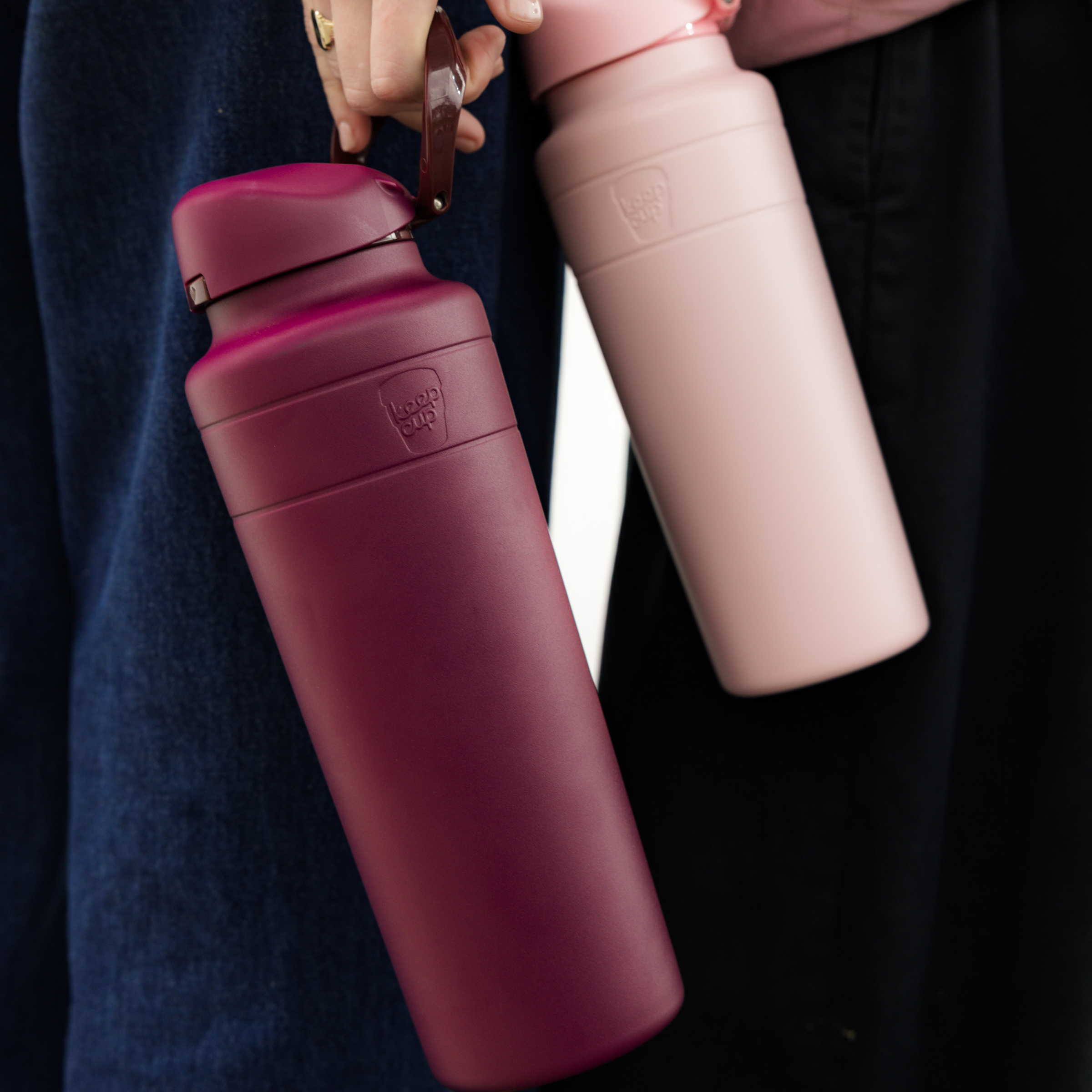 A hand holds the KeepCup Ora Bottle 32oz in Mulberry by its carry loop, showing the deep burgundy matte finish, while a second person beside them carries a pale pink Ora Bottle tucked under their arm.