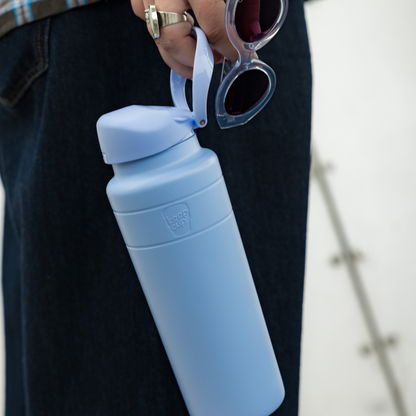 A hand holding the KeepCup Ora Bottle 24oz in Orchid by its carrying loop, alongside a pair of sunglasses, against a backdrop of dark denim jeans, showcasing the bottle's matte light blue finish and compact design.