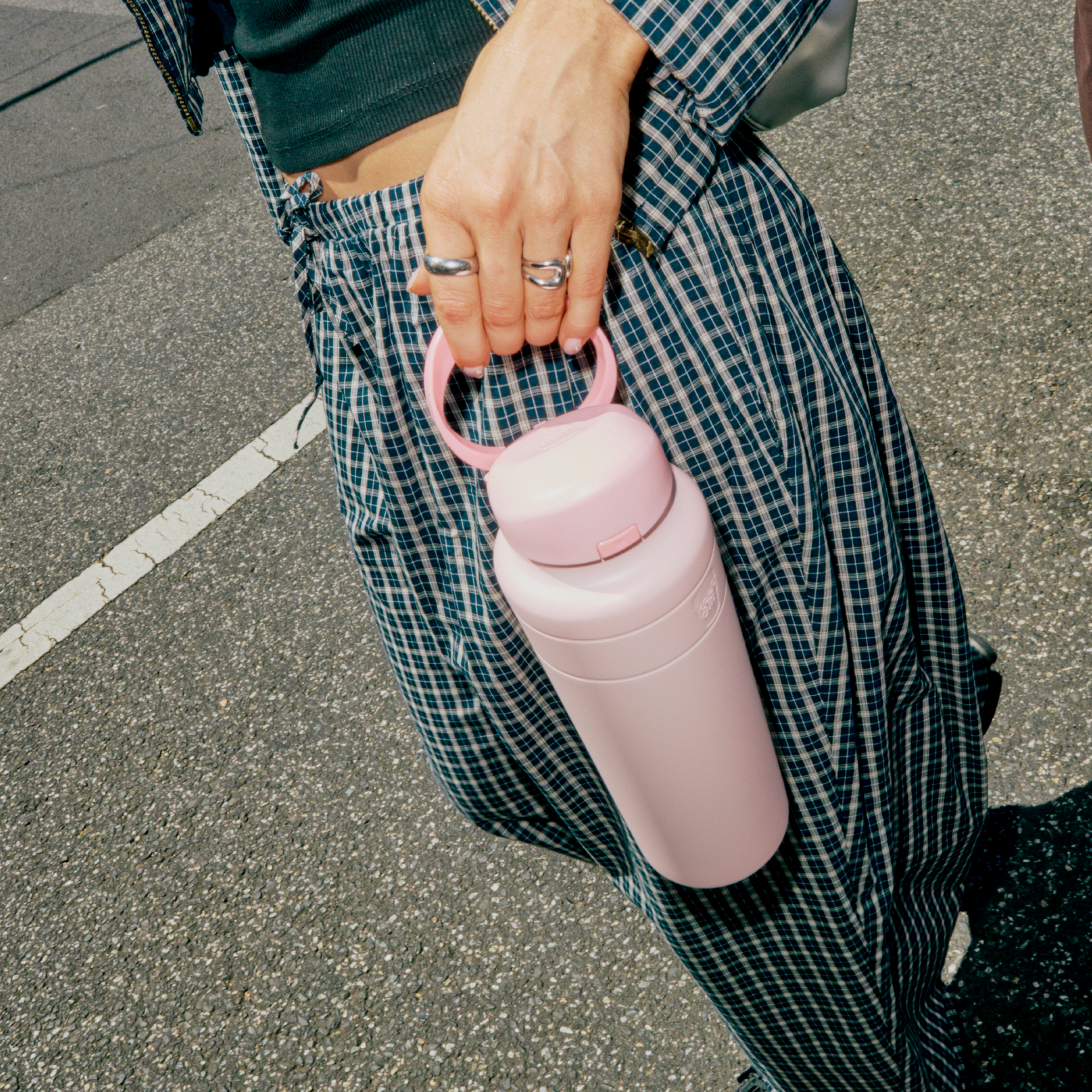 A person in blue-green plaid pants and a dark cropped top carries the Ora Bottle 24oz in Pashmak Pink by its handle while walking across a parking lot, with silver rings visible on their fingers.