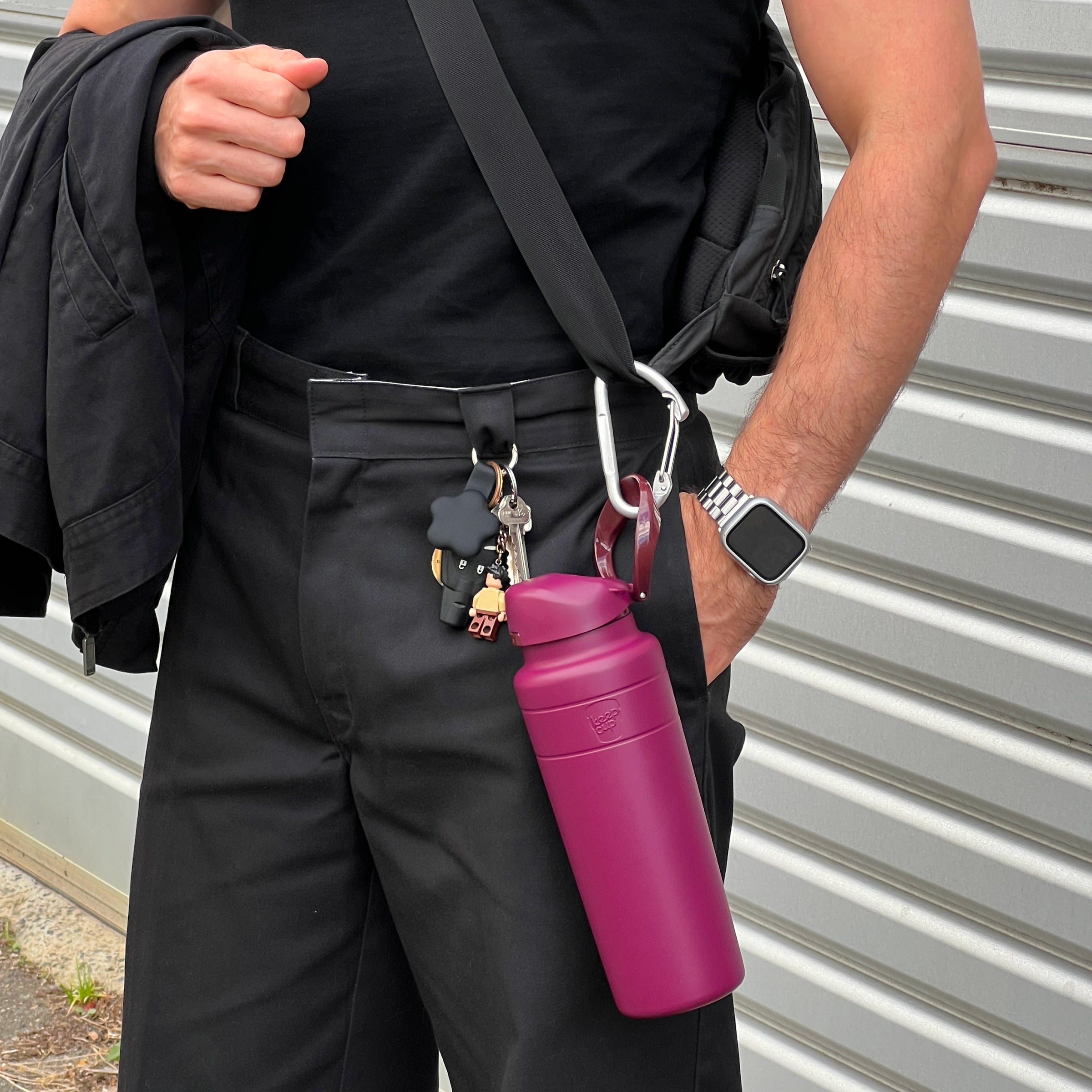 A person dressed in all black holds their backpack strap while a KeepCup Ora Bottle 32oz in Mulberry hangs from a silver carabiner attached to the bag, showing the bottle's built-in carry loop and deep magenta color against their dark outfit.