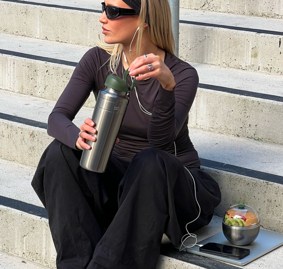 A woman sits on concrete outdoor steps wearing a fitted dark plum long-sleeve athletic top and black pants, holding an Ora insulated water bottle in Nitro Pine. She has white earphones in, insulated Go Bowl Luxe filled with fruit and a phone sit beside her on the steps.