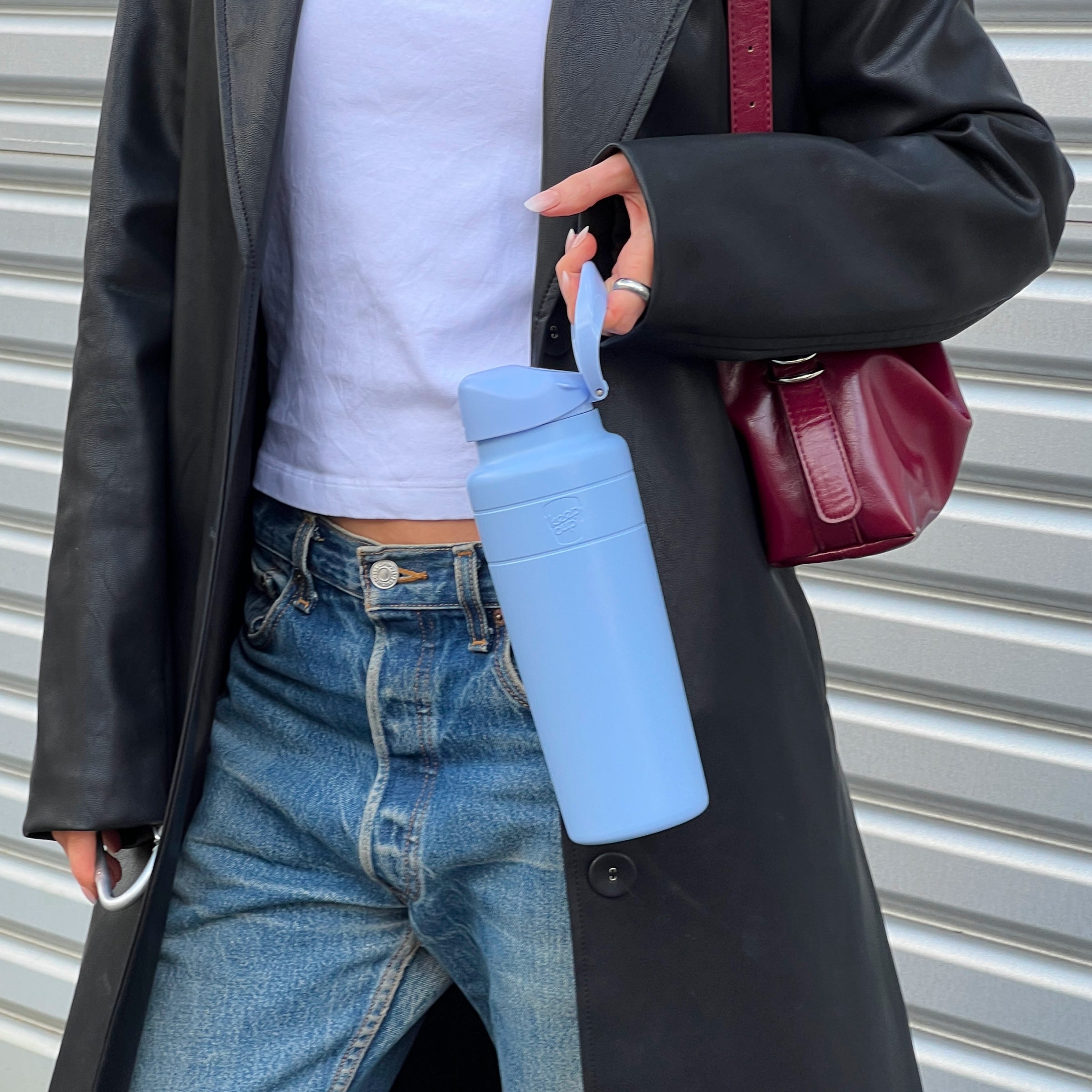 A person in a black leather coat, white crop top, and blue jeans holds a KeepCup Ora Bottle 24oz in Orchid color by its carry loop, showcasing the light blue insulated bottle with flip-top lid against an urban corrugated metal backdrop.