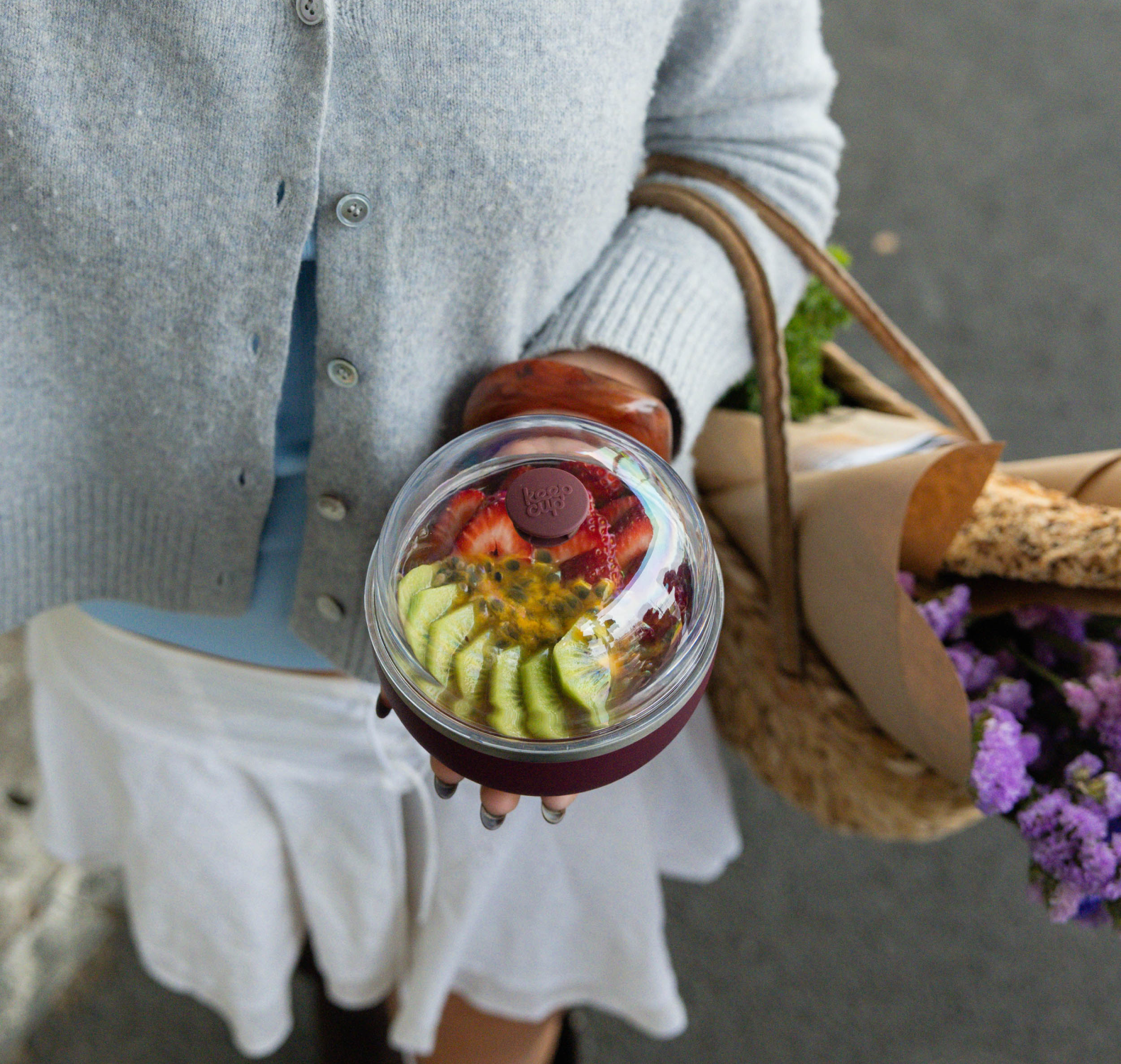 A person in a gray cardigan and white skirt holds a Go Bowl Luxe filled with strawberries, kiwi slices, and passion fruit, with a woven straw bag and purple flowers beside them.