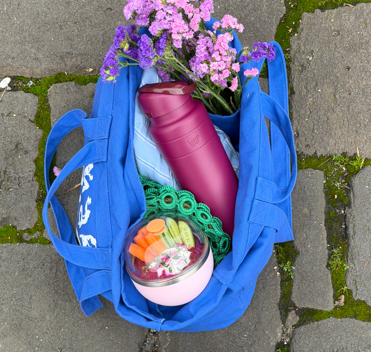 Blue bag with an Ora Bottle in Mulberry and Go Bowl Luxe in Pashmak Pink wiith dip and flowers on a stone pavement.